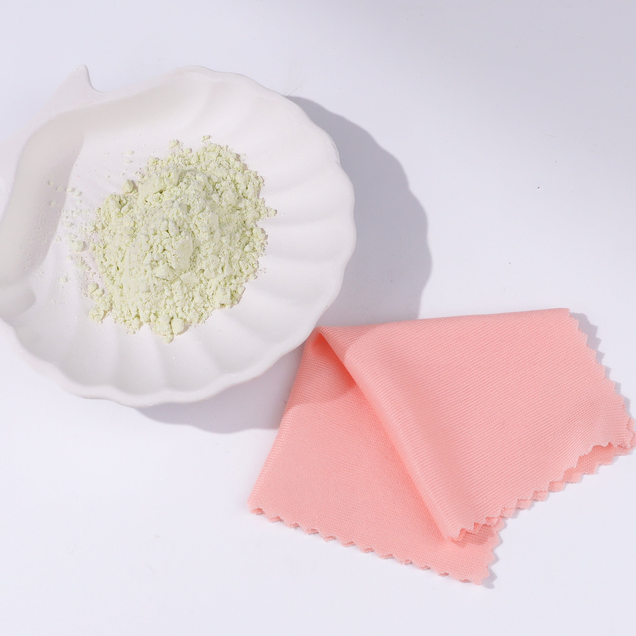 White bowl with green powder next to a pink cloth on a white background